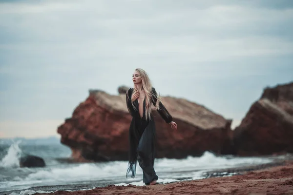 Giovane donna in lungo abito nero trasparente tra spiaggia e mare . — Foto Stock