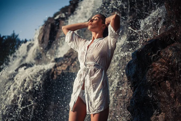 Jonge vrouw in wit shirt en bikini staat in de buurt van de waterval. — Stockfoto