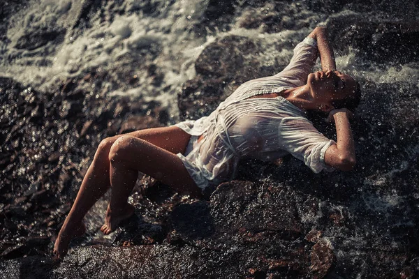 Jonge vrouw in wit shirt en bikini ligt op rots in de buurt van waterfal — Stockfoto