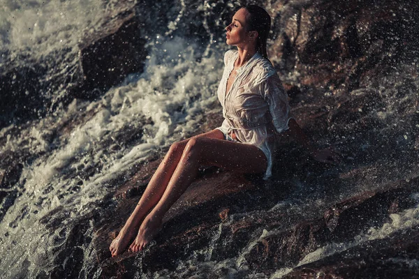 水の流れに岩の上に座っている白いシャツとビキニの若い女性 — ストック写真