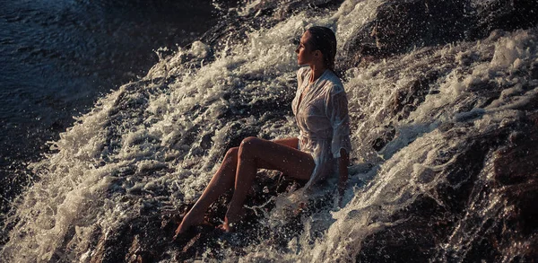 Jovem mulher de camisa branca e biquíni senta-se na rocha em fluxo de água — Fotografia de Stock