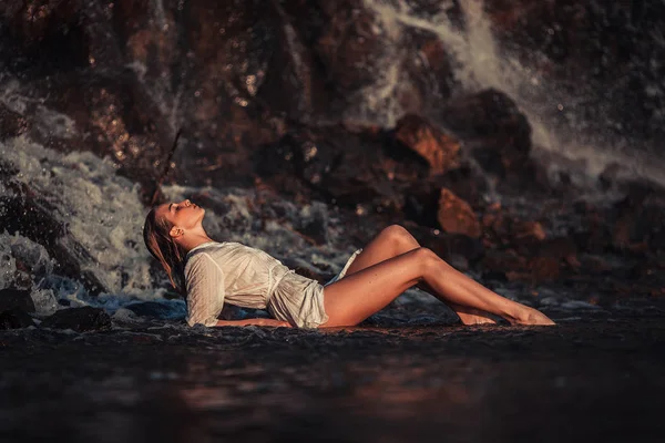 Young woman in white shirt and bikini lies on rock near waterfal — Stock Photo, Image
