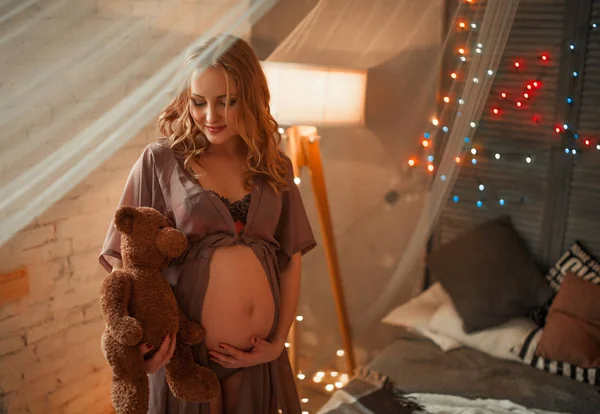 妊娠中の女性のライトの中でベッドの上に座っているし、手で彼女の腹を抱擁. — ストック写真
