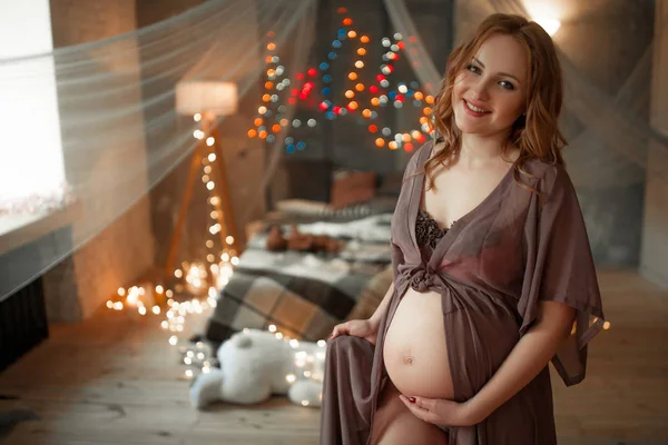 Zwangere vrouw zit op bed onder lichten en haar buik met handen hugs. — Stockfoto