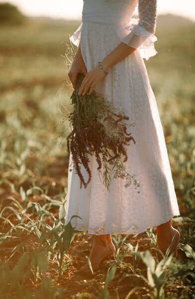 Young woman in long white lace dress on cornfield. She stands barefoot with bouquet of wild flowers in her hands.