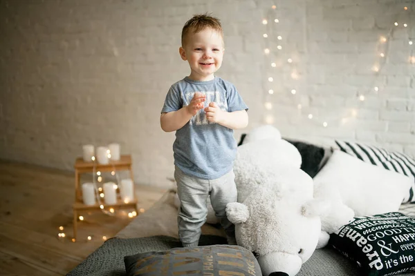 テディベアと笑顔の横にあるベッドの上に幼児が立っています。. — ストック写真