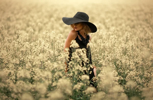 Jeune femme en robe noire et chapeau parmi la prairie fleurie . — Photo