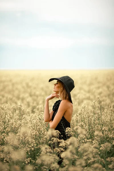 Ung kvinna i svart klänning och hatt bland blommande äng. — Stockfoto