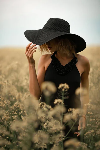 Jovem mulher em vestido preto e chapéu entre o prado florido . — Fotografia de Stock