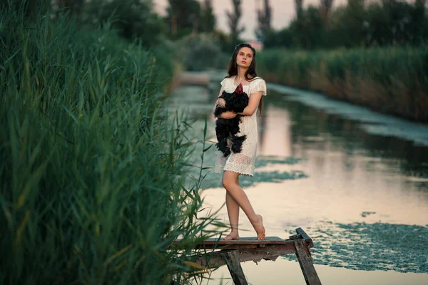 Jovem mulher rural fica na ponte de madeira sobre o lago com galo nas mãos . — Fotografia de Stock