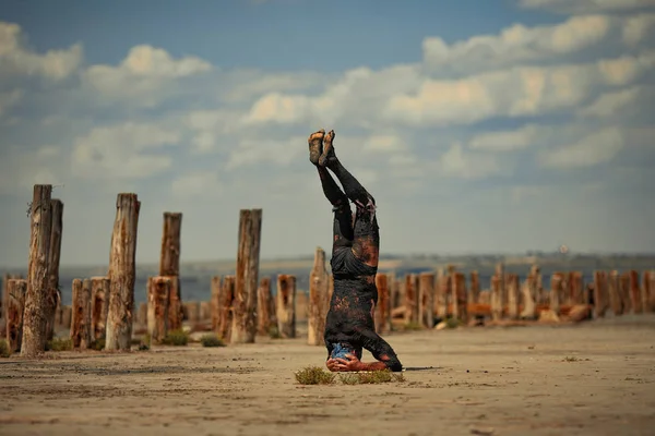 Young man smeared with therapeutic mud and stands on head on beach background. — Stock Photo, Image