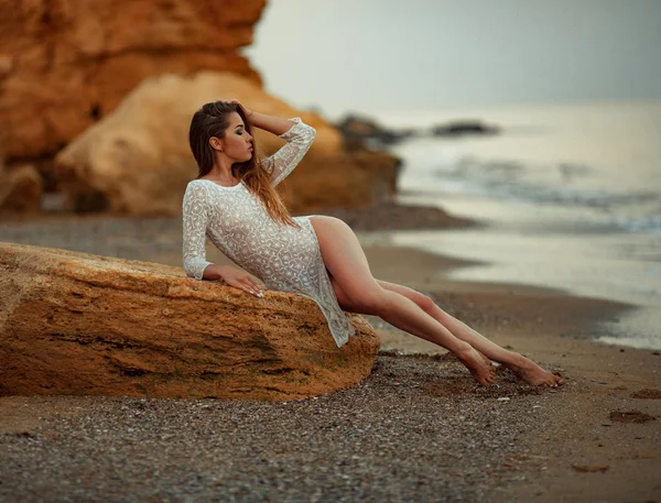 Giovane donna in abito di pizzo bianco si trova su roccia sulla spiaggia di ghiaia in riva al mare . — Foto Stock