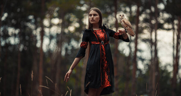 Girl in red and black dress stands with owl on her hand in forest and looks away.
