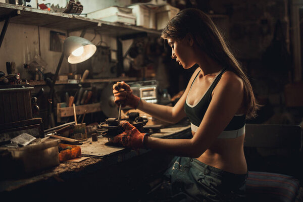 Girl worker in t-shirt sits in workshop among tools and unscrews Stock Photo