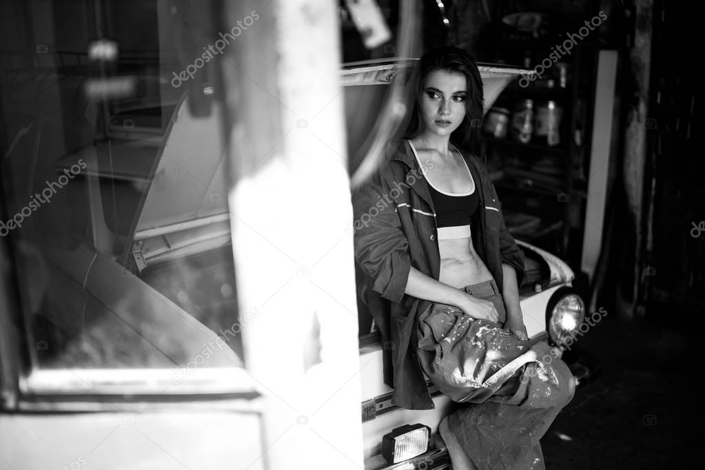 Girl worker in overalls stands in workshop near opened capote of