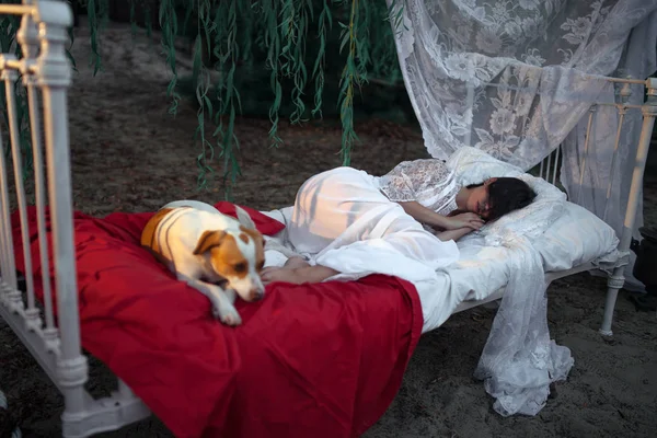 Jeune femme avec chien se trouve sur le lit avec literie et baldaquin près de l'arbre et dort . — Photo