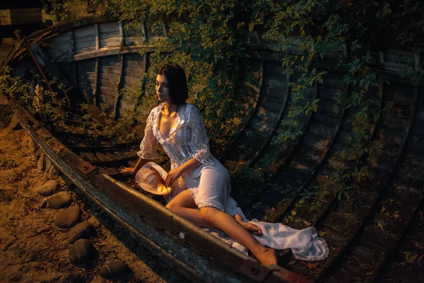 Jeune femme sexy est assis dans un bateau abandonné le soir. Elle est éclairée par le coucher du soleil . — Photo