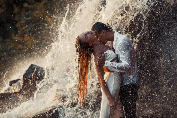 Young enamored couple hugs and kisses under spray of waterfall. — Stock Photo, Image