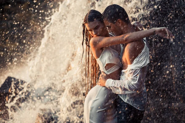 Young enamored couple hugs under spray of waterfall. — Stock Photo, Image