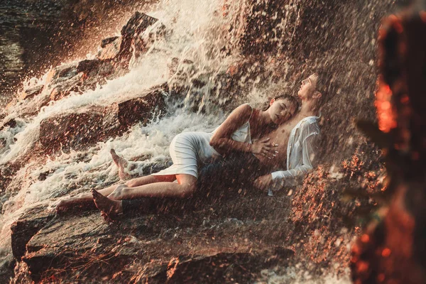 Νεαρό ζευγάρι ερωτευμένων καθώς κοιτάζονται βρίσκεται πάνω σε βράχο και αγκαλιές υπό ψεκασμό του καταρράκτη. — Φωτογραφία Αρχείου