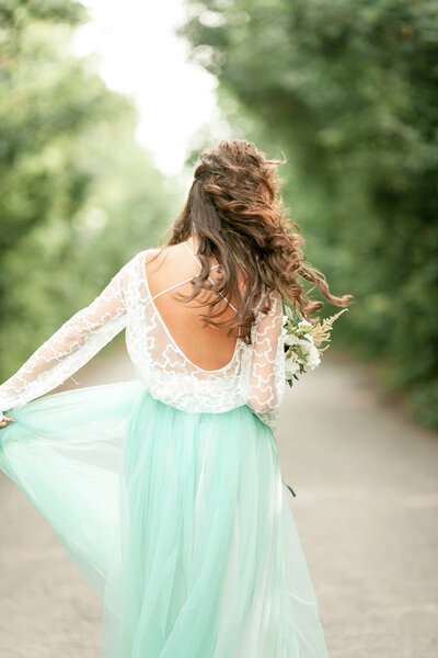 Bride in beautiful wedding dress goes on forest road with bouquet in hands, back view.