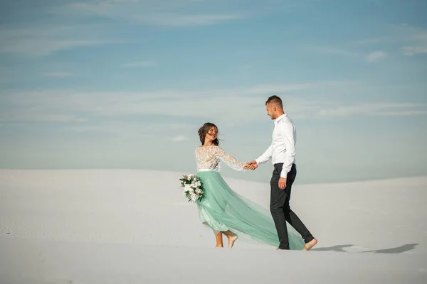 Gelin ve damat el ele, gülümseme ve çölün ortasında yalınayak yürümek. — Stok fotoğraf