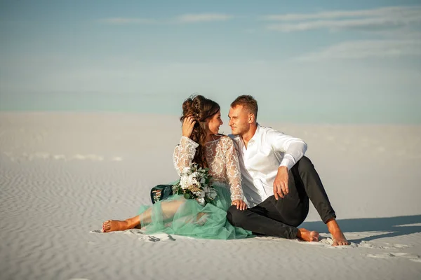 Menyasszony és a vőlegény ülni mezítláb a fehér homok a sivatagban, ölelés és a lo — Stock Fotó