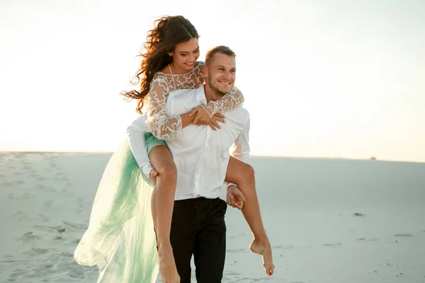El novio lleva a la novia en su espalda a través de la arena en el desierto. De cerca. . —  Fotos de Stock