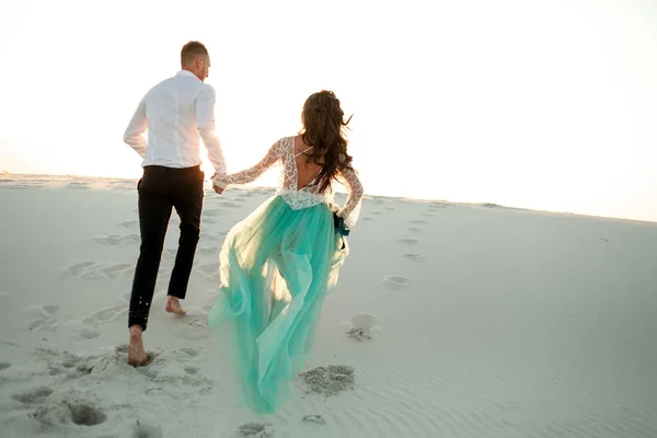 Bruden och brudgummen håller händer och köra på sand i öknen. Bakifrån. — Stockfoto