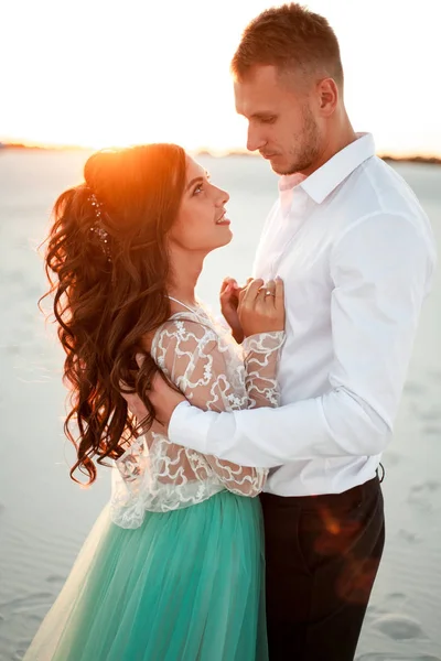 Braut und Bräutigam stehen bei Sonnenuntergang in der Wüste, lächeln und umarmen sich. cl — Stockfoto