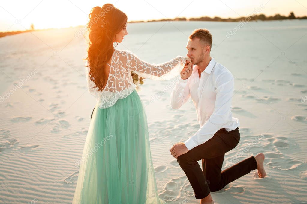Groom stands barefoot before bride on one knee in desert on suns
