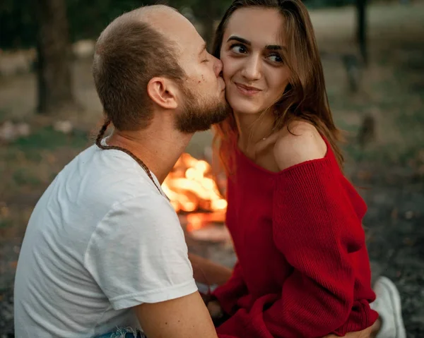 Pareja encantada se sienta y besa en el picnic en el bosque en el fondo de la llama de la hoguera . — Foto de Stock