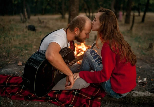 Verliefd paar zit op picknick in het bos met gitaar op achtergrond van vreugdevuur vlam. — Stockfoto
