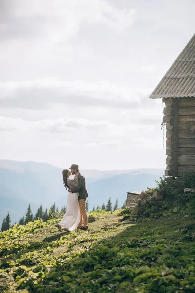 Newlyweds stand near wooden hut and hug in mountains. — Stock Photo, Image