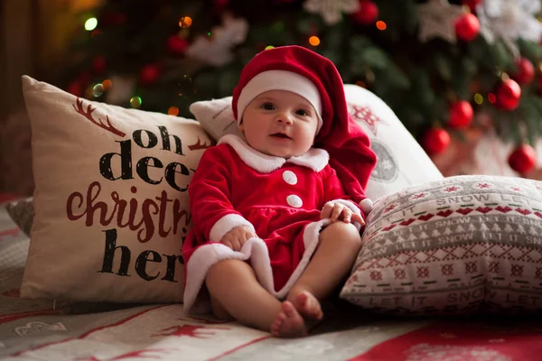 Niña en traje de Santa Claus se sienta y sonríe entre almohadas contra el fondo de las decoraciones de Navidad . —  Fotos de Stock
