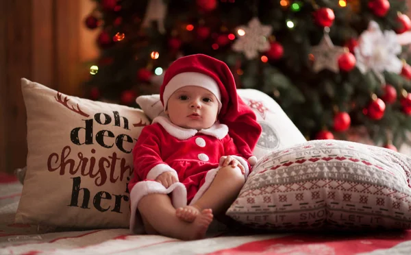 Niña en traje de Santa Claus se sienta entre almohadas contra el fondo de las decoraciones de Navidad . —  Fotos de Stock