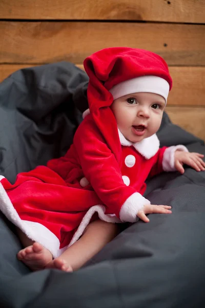 Sonriente niña en traje de Santa Claus se sienta entre almohada contra fondo de tablones de madera . —  Fotos de Stock
