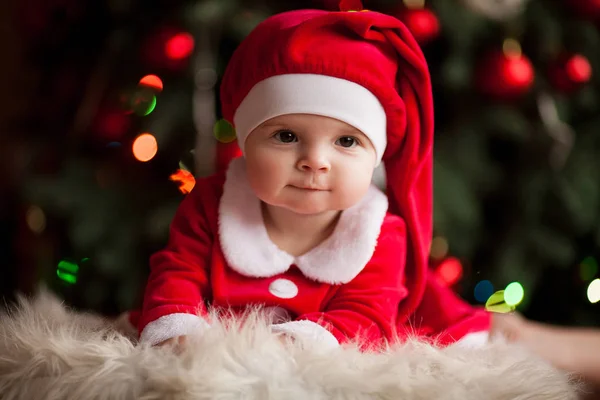 Allegro bambino ragazza in costume di Babbo Natale si trova sul tappeto di pelliccia sullo sfondo di luci di Natale . — Foto Stock