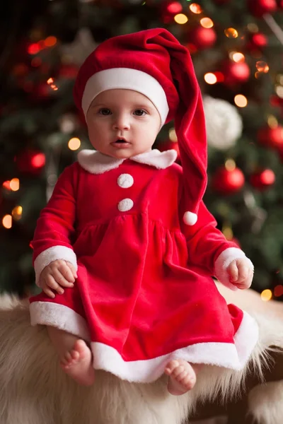 Niña en traje de Santa Claus se sienta en la alfombra de piel contra el fondo de las luces de Navidad . —  Fotos de Stock