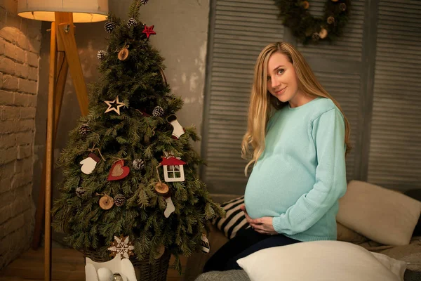 Mulher grávida senta-se antes da árvore de Natal e suporta a barriga . — Fotografia de Stock