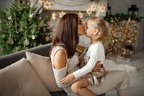 母はソファーに座っているし、クリスマスの装飾の背景と彼女の小さな娘にキス. — ストック写真