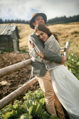 Yeni evliler standı, gülümseme ve eski ahşap kulübe ve Karpat Dağları'nda çit arka hug.