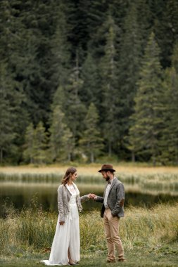 Gelin ve damat ayakta ve orman göl kenarında elele.
