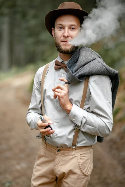 Молодий чоловік в стильному одязі стоїть і курить електронну сигарету . — стокове фото