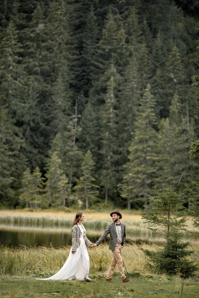 Nevěsta a ženich jsou děje a drželi se za ruce v blízkosti forest lake. — Stock fotografie