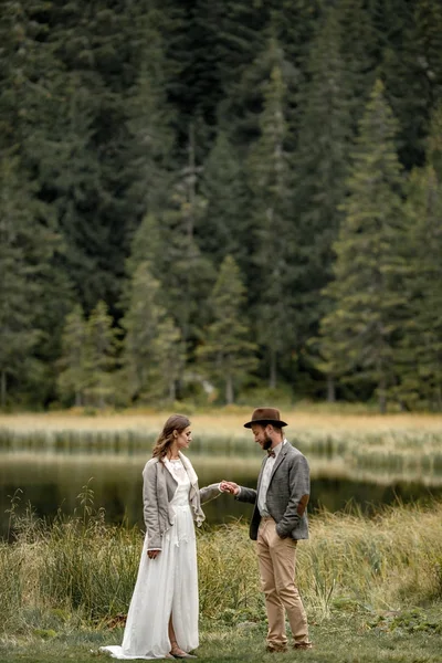 Gelin ve damat ayakta ve orman göl kenarında elele.