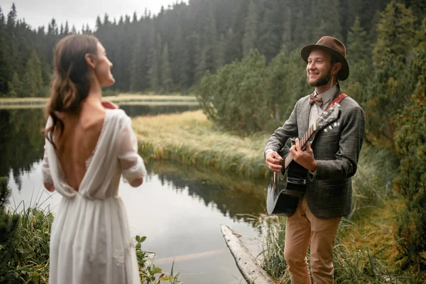 Groom joue de la guitare et chante une chanson pour la mariée près du lac forestier . — Photo