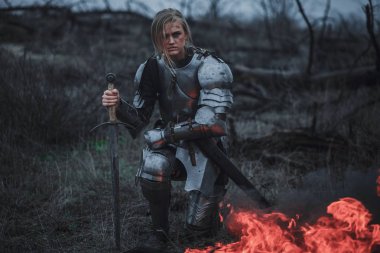 Jeanne d'Arc zırh ve onun elinde kılıçla görüntüsünü kız ateş ve duman arka planı diz çöker..