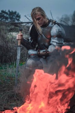 Jeanne d'Arc zırh ve onun elinde kılıçla görüntüsünü kız ateş ve duman arka planı diz çöker..