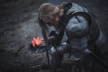 Kız Jeanne d'Arc görüntü ile kılıç ve zırh ve ateş ve duman çerçevede onun elinde diz çöker.
